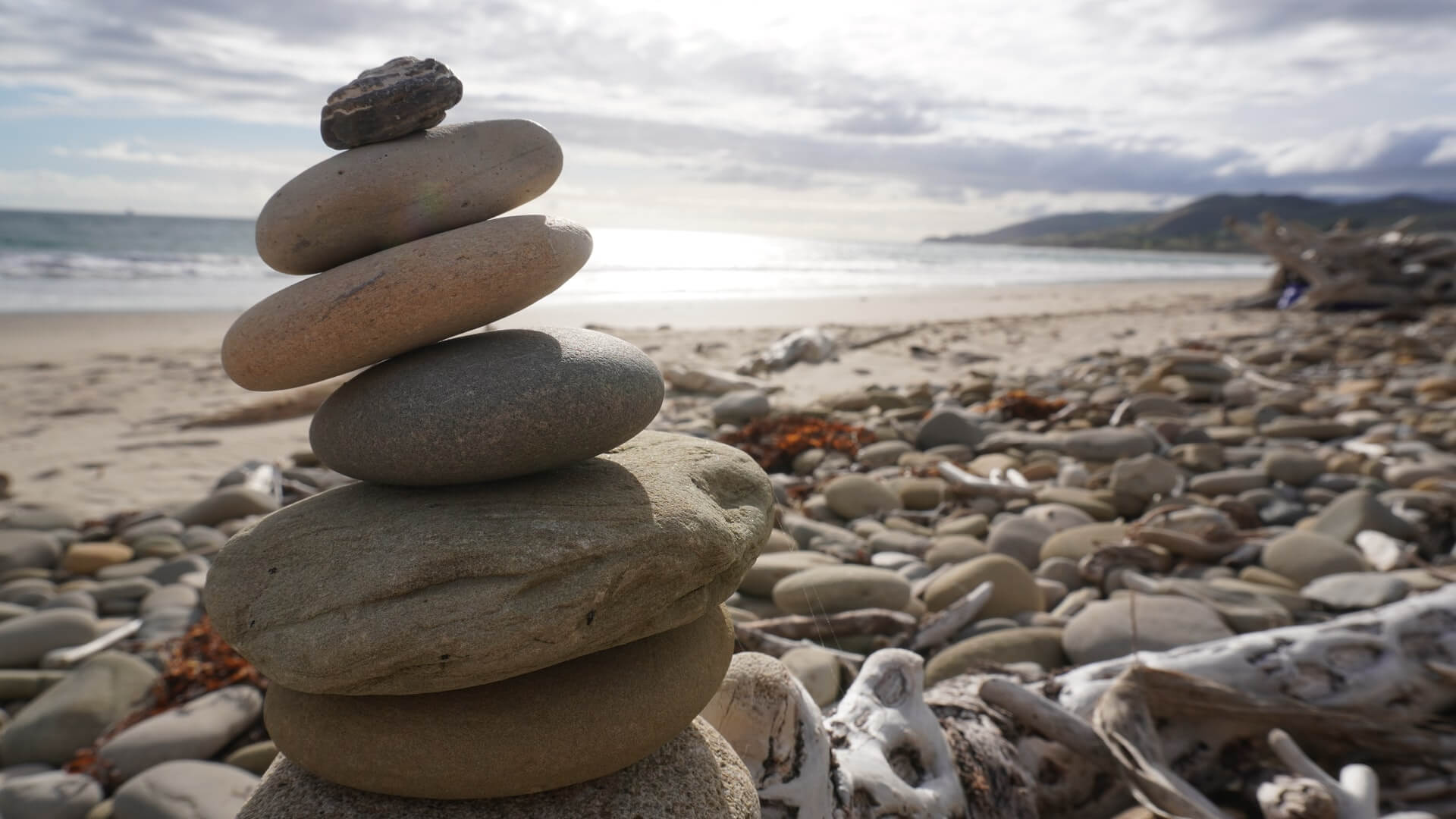 A group of gray and tan rocks stacked on a rocky, sandy beach (Paradise Cove Beach, California, USA). The sun is shining with mountains in the background.