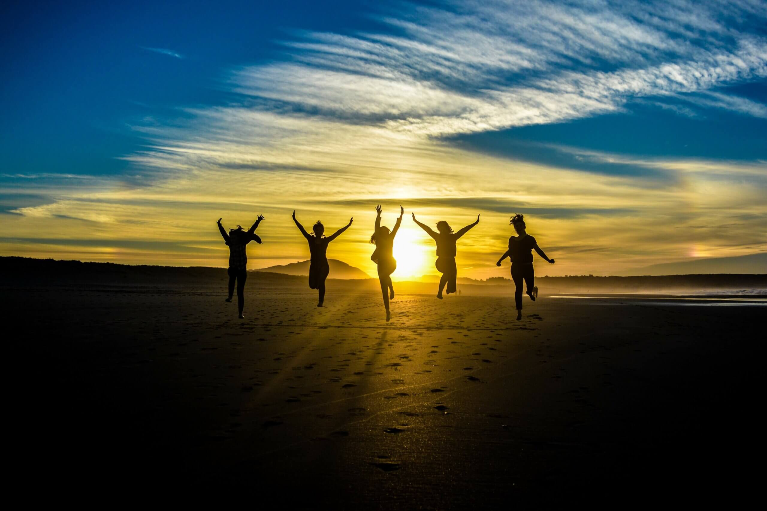 5 women jumping on a beach in Spain. They are dark silhouettes with a blue sky and setting sun behind them.