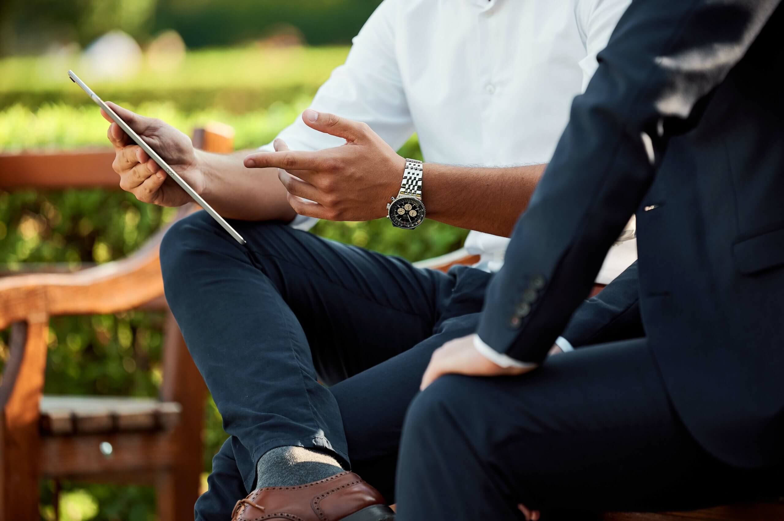 Close up of two people sitting on a park bench. Person in a navy suit in foreground. Person in a white shirt and navy pants sitting next to them, pointing at a tablet screen.